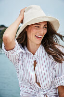 A brunette woman by the sea wearing a hat and a striped shirt-blouse