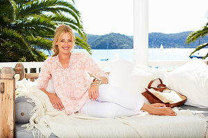 A blonde woman lying on a daybed wearing a tunic blouse and white trousers