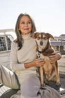 A grey-haired woman wearing a turtleneck jumper and light trousers with a dog