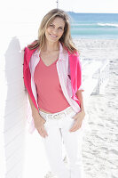A long-haired woman on the beach wearing a long blouse with a pink jumper over her shoulders