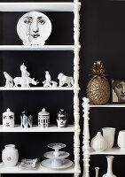 Porzellanfiguren und Fornasetti-Deko im Regal vor schwarzer Wand