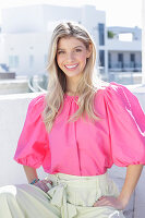 A young blonde woman wearing a pink top with puffed sleeves and white summer trousers