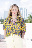 A young blonde woman wearing a black-and-yellow patterned blouse and a pair of light trousers
