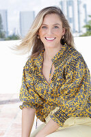 A young blonde woman wearing a black-and-yellow patterned blouse and a pair of light trousers