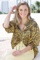 A young blonde woman wearing a black-and-yellow patterned blouse and a pair of light trousers