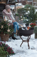 Frau trinkt Tee auf der verschneiten Terrasse mit Feuerschale und Weihnachtsbaum