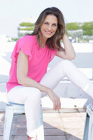 A long-haired woman wearing a pink summer top and white trousers