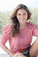 A young, long-haired woman wearing a pink blouse and shorts sitting in the sand