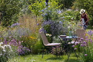 Gemütlicher Sitzplatz am Staudenbeet mit Duftnessel 'Blue Fortune' 'Apache Sunset', Mannstreu 'Glitter Blue', Färberkamille, Gräser, Scheinsonnenhut, Petunie, Berglauch, Bleiwurz und Spinnenblume, Frau bringt Krug mit Wasser, Gläser und Blumenstrauß auf dem Tisch