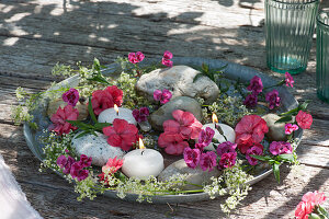 Schale mit Nelkenblüten, Labkraut, Schwimmkerzen und groben Kieselsteinen im Wasser