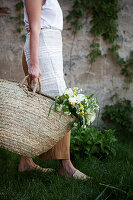 Woman carrying white bouquet of peonies, viburnum, feverfew and grasses in basket bag