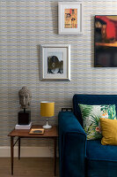 Sofa with blue velvet cover and cushions, side table with Buddha and patterned wallpaper with photo and poster