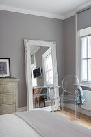 Large mirror with wooden frame leaning against grey wall and designer chair in bedroom