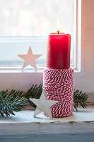 Red candle on a spool of thread, fir branch and wooden stars on a windowsill