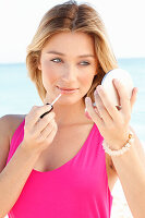 Young blonde woman in a pink dress cares for her lips with lip gloss