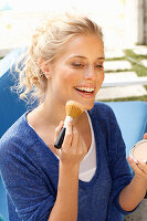 Blonde Frau in blauem Shirt trägt sich Make-up mit Pinsel auf