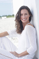 Fröhliche, langhaarige Frau im weißen Pullover und weißer Hose am Strand