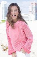 A laughing, long-haired woman wearing a pink jumper and white trousers
