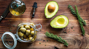 Avocado, eingelegte grüne Oliven, Olivenöl und Rosmarin auf Holztisch