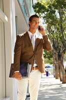 A young businessman wearing a brown jacket and light-coloured trousers talking on the phone in the street