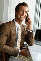 Junger Geschäftsmann mit braunem Sakko sitzt im Büro und telefoniert