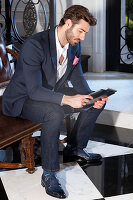 Young man with a beard in a jacket and jeans sitting on a leather sofa, holding a tablet