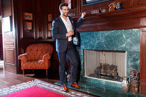 Young man with a beard in a jacket and jeans is standing in front of a marble fireplace