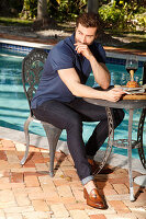 Young man with a beard in a blue polo shirt and jeans is sitting at the table by the pool