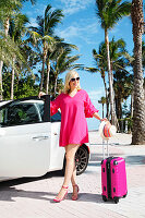 Junge blonde Frau in pinkem Sommerkleid mit farblich passendem Trolley am Wagen