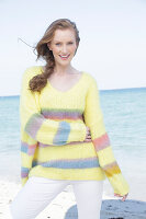 Young, blonde woman in a yellow jumper with coloured stripes and white trousers at the seaside