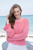 Young, blonde woman in a pink jumper and white trousers by the sea