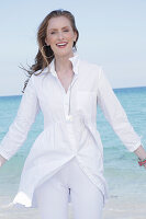 Young, blonde woman in a white blouse dress and white trousers by the sea
