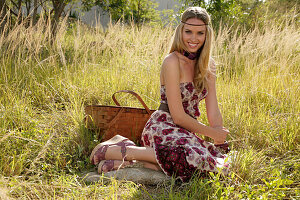 Blonde woman in off-the-shoulder summer dress with picnic basket on the meadow