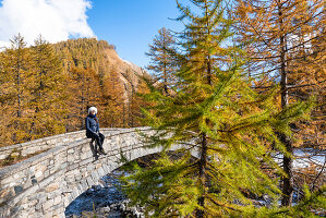 Ein Mädchen sitzt auf der Brücke des Flusses Cairasca umrahmt von gelben Lärchen im Herbst, Alpe Veglia, Val Cairasca Tal, Divedro Tal, Ossola Tal, Varzo, Piemont, Italien, Europa