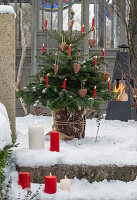 Geschmückter Christbaum mit Wachskerzen auf verschneiter Terrasse und Winterfeuer