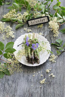 Bund grüner Spargel auf weißem Teller dekoriert, mit Storchenschnabel (Geranium), Holunderblüten (Sambucus Nigra) und Namensschild