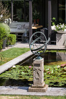 Garden with sundial sculpture and water lily pond in front of modern terrace