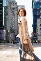 Junge Frau mit Brille und Tasche in hellem Mantel auf der Straße mit Hochhäusern