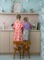 Two children on chairs in a pastel-colored kitchen with floral wallpaper