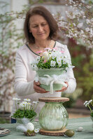 Woman building tiered flower pot made of horned violets, daisies and glass vase, Easter eggs in small moss nests with feathers on table