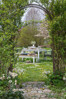 Gedeckter Tisch im Garten für Osterfrühstück mit Osternest und gefärbten Eiern, Blumenstrauß, Korb mit Eiern in der Wiese, Torbogen durch Kletterpflanzen
