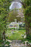 Gedeckter Tisch im Garten für Osterfrühstück mit Osternest und gefärbten Eiern, Blumenstrauß, Hund in der Wiese, Torbogen durch Kletterpflanzen