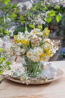 Blumenstrauß aus Schlüsselblume (Primula veris), Felsenbirne (Amelanchier), Narzissen 'Bridal Crown' (Narcissus) in Glasvase , Osterhasenfigur und Eierschale auf Gartentisch