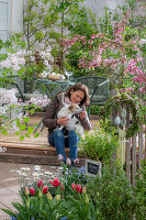 Zwergflieder 'Palibin', Blutpflaume 'Nigra', Tulpen, Marokkomargerite, Vergissmeinnicht und Kräuter im Blumenbeet, Frau mit Hund vor gedecktem Tisch auf Terrasse