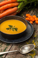 A bowl of creamy carrot soup garnished with fresh herbs, surrounded by whole and sliced carrots on a wooden table.