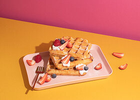 From above tasty sweet waffles topped with berries fruits sauce and cream served on pink tray on colorful table background in light kitchen