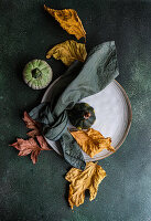 Draufsicht auf einen herbstlich gedeckten Tisch mit Serviette, Blättern und Kürbissen auf einem Teller vor einer dunklen Fläche