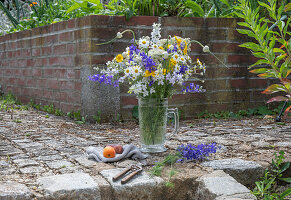 Blumenstrauß aus Rittersporn (Delphinium), Färberkamille (Anthemis tinctoria), Etagenzwiebel, Große Knorpelmöhre, auf Gartenmauer im Bierkrug