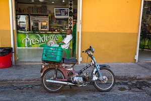 A delivery motorycle in front of a neighborhood minimart in the Dominican Republic. A man waits for a delivery order.