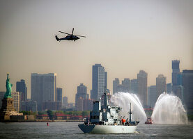 HMS Scott, schwebender Hubschrauber und Schlepper, die in der Nähe der Freiheitsstatue Wasser versprühen, mit Ellis Island und der Skyline von Jersey City, New Jersey, im Hintergrund, Hafen von New York, New York, USA